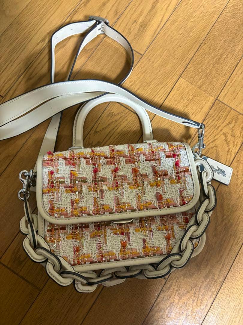 Front view of the COACH tweed shoulder bag with cream leather trim, chain-link strap, and multicolored tweed front placed on a wooden floor.