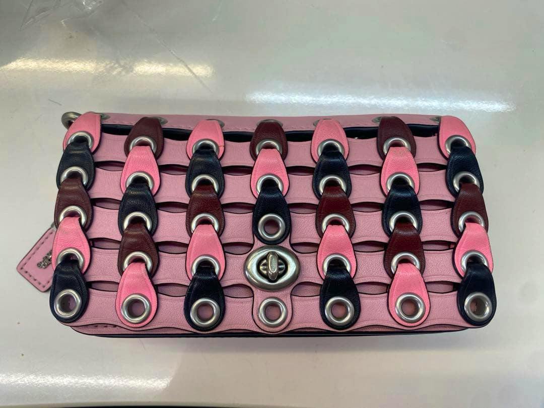 Front of a Coach ultra-mini pochette wallet in pink cow leather with interlaced multicolored leather tabs, silver grommets, and a turn-lock closure.