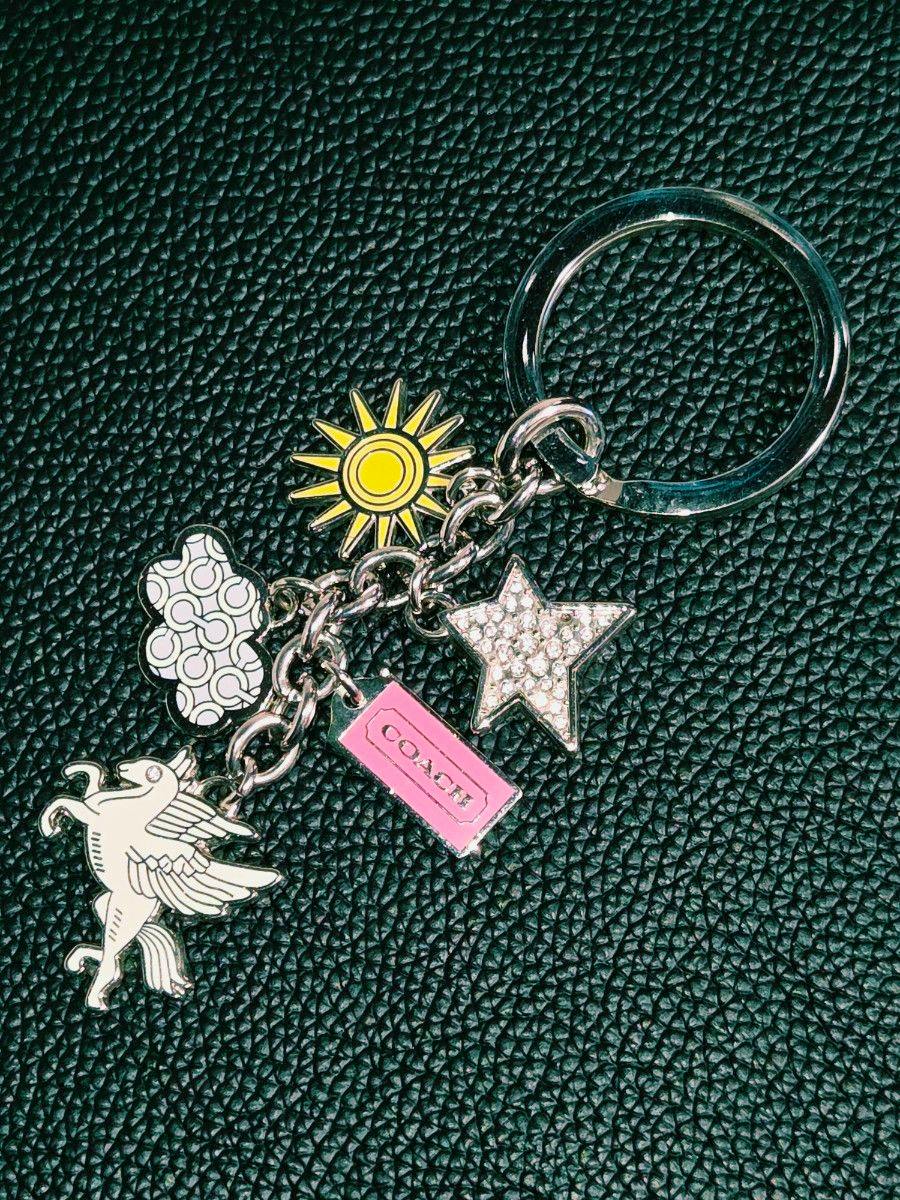 Silver Coach key ring bag charm showing a unicorn, patterned cloud, yellow sun, rhinestone star and a pink COACH tag on a dark textured background.