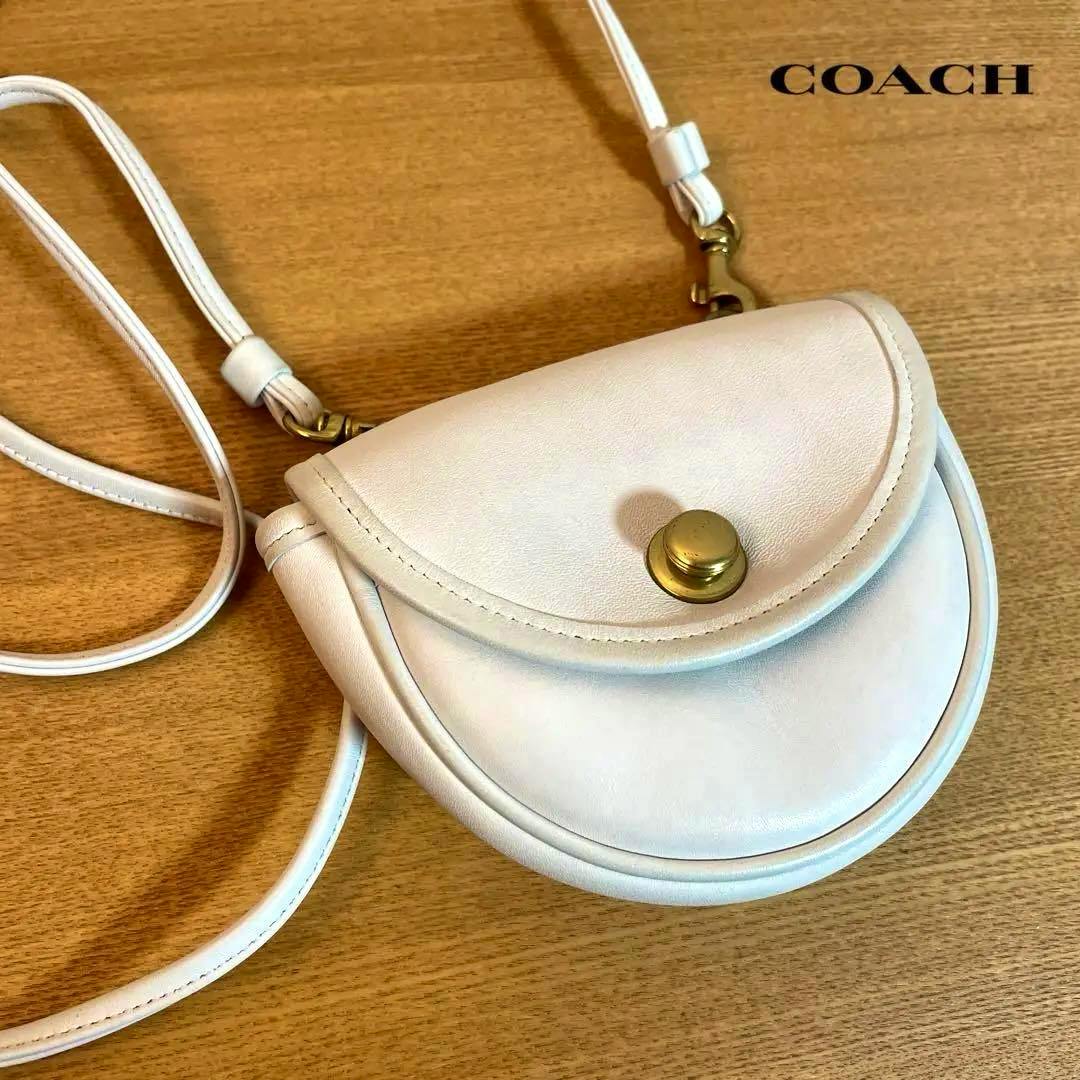 White COACH cow-leather mini shoulder bag with a round flap and brass snap closure displayed on a wooden surface.