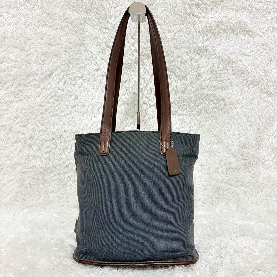Front view of a COACH twill canvas tote bag with brown leather handles and a leather name tag on a light textured background.