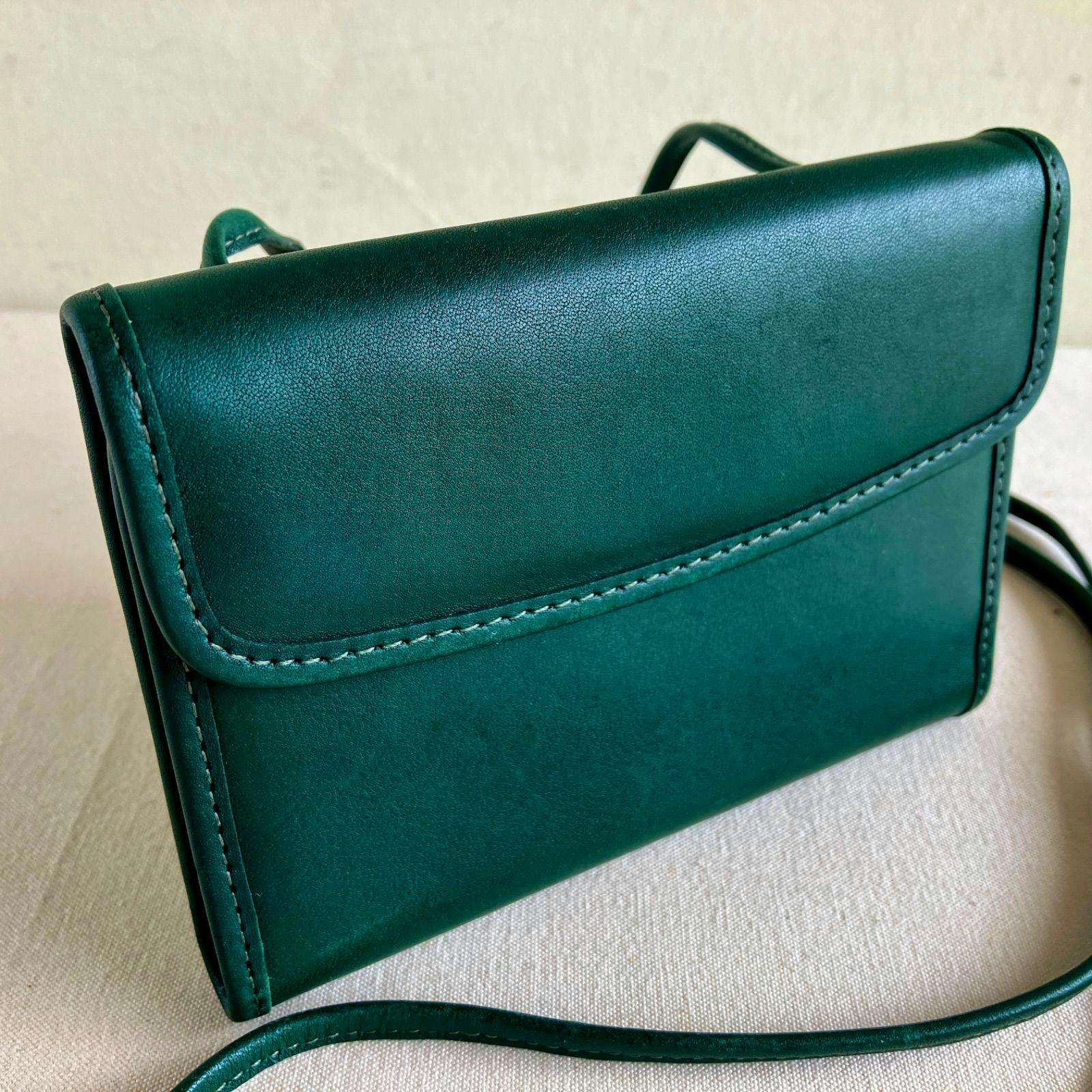 Close-up of a bottle-green Coach vintage envelope swing wallet crossbody showing the front flap, stitching, and thin leather strap on a light fabric surface.