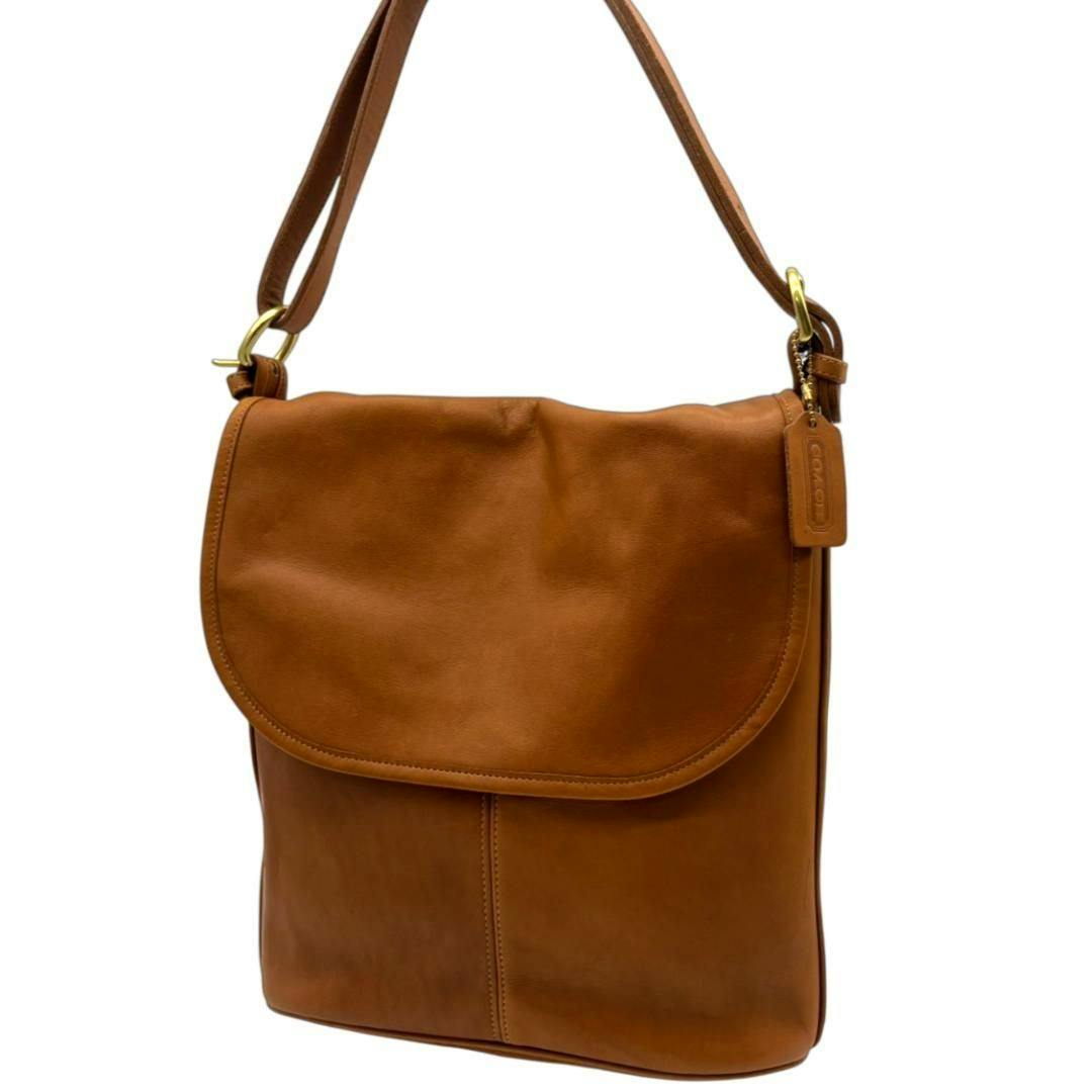 Brown all-leather vertical Coach shoulder flap bag hanging by its strap, showing a leather logo tag and gold-tone hardware.