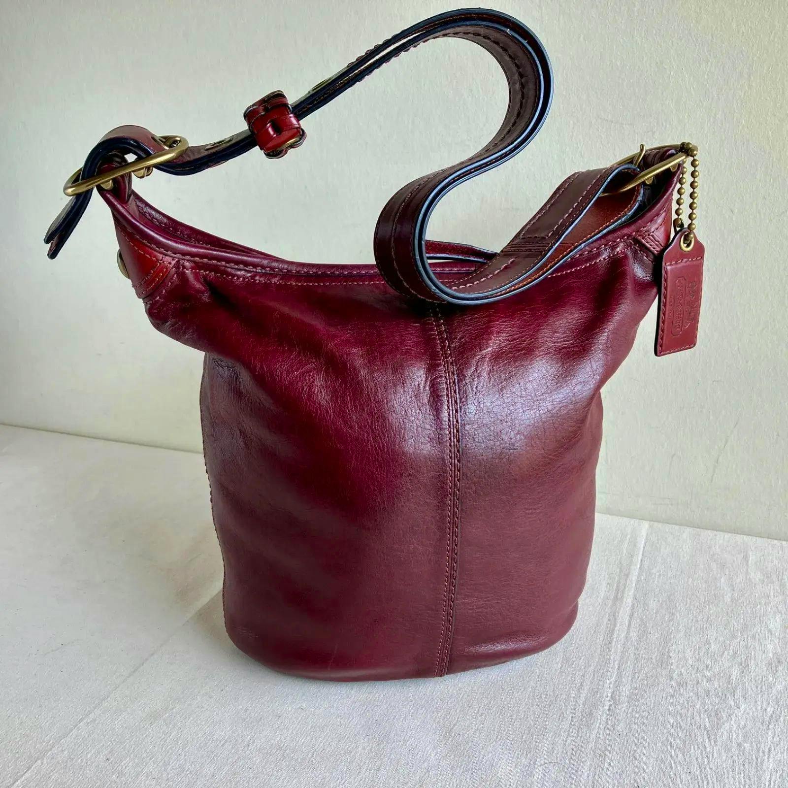 Wine burgundy leather Coach bucket-style duffle shoulder bag shown upright with its strap and leather hang tag.