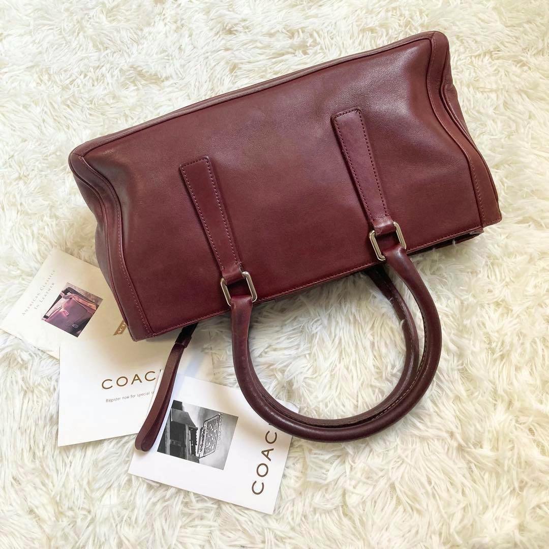 Brown leather COACH mini Boston handbag shown lying on a white faux-fur surface with COACH booklets and the bag handles visible.