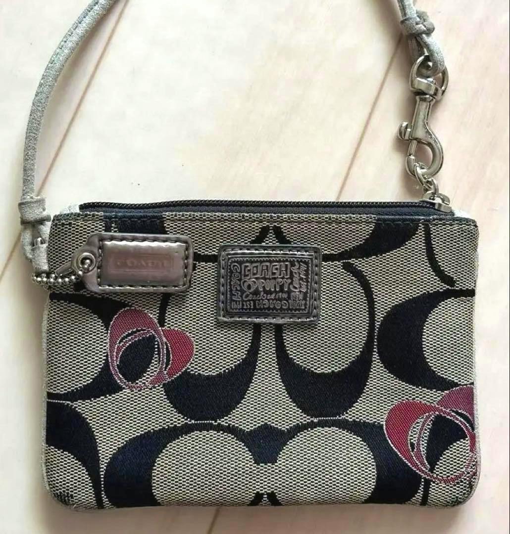 A Coach canvas coin case with signature C pattern, pink heart accents, and a wrist strap placed on a light wood floor.