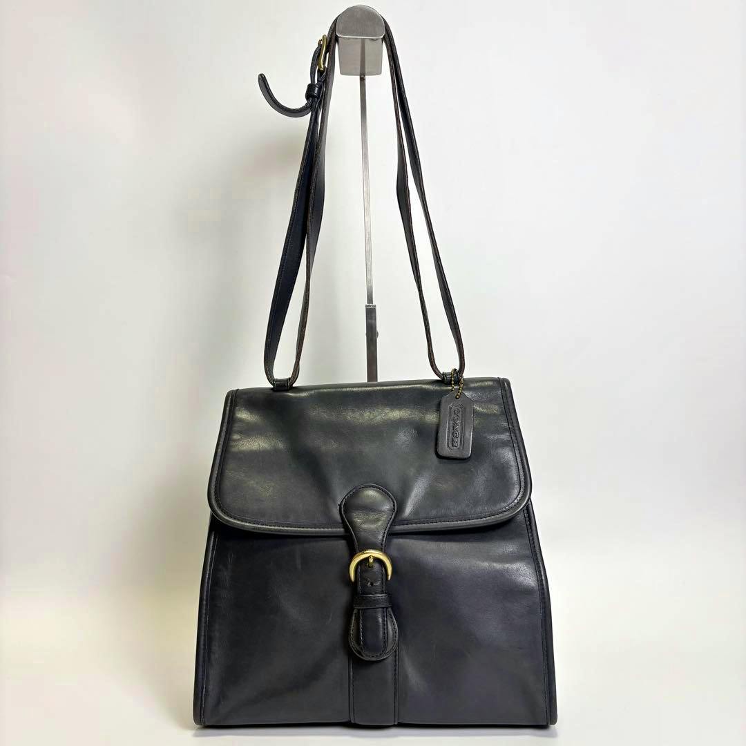 Black Coach Kelly leather shoulder bag with flap and gold-tone buckle hanging on a stand against a white background.