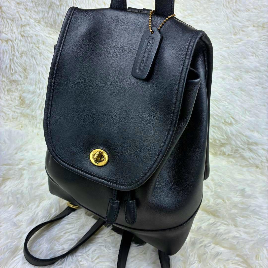 Black COACH leather backpack shown from the front/angled view with flap, gold turn-lock closure, and COACH leather hang tag on a white textured background.