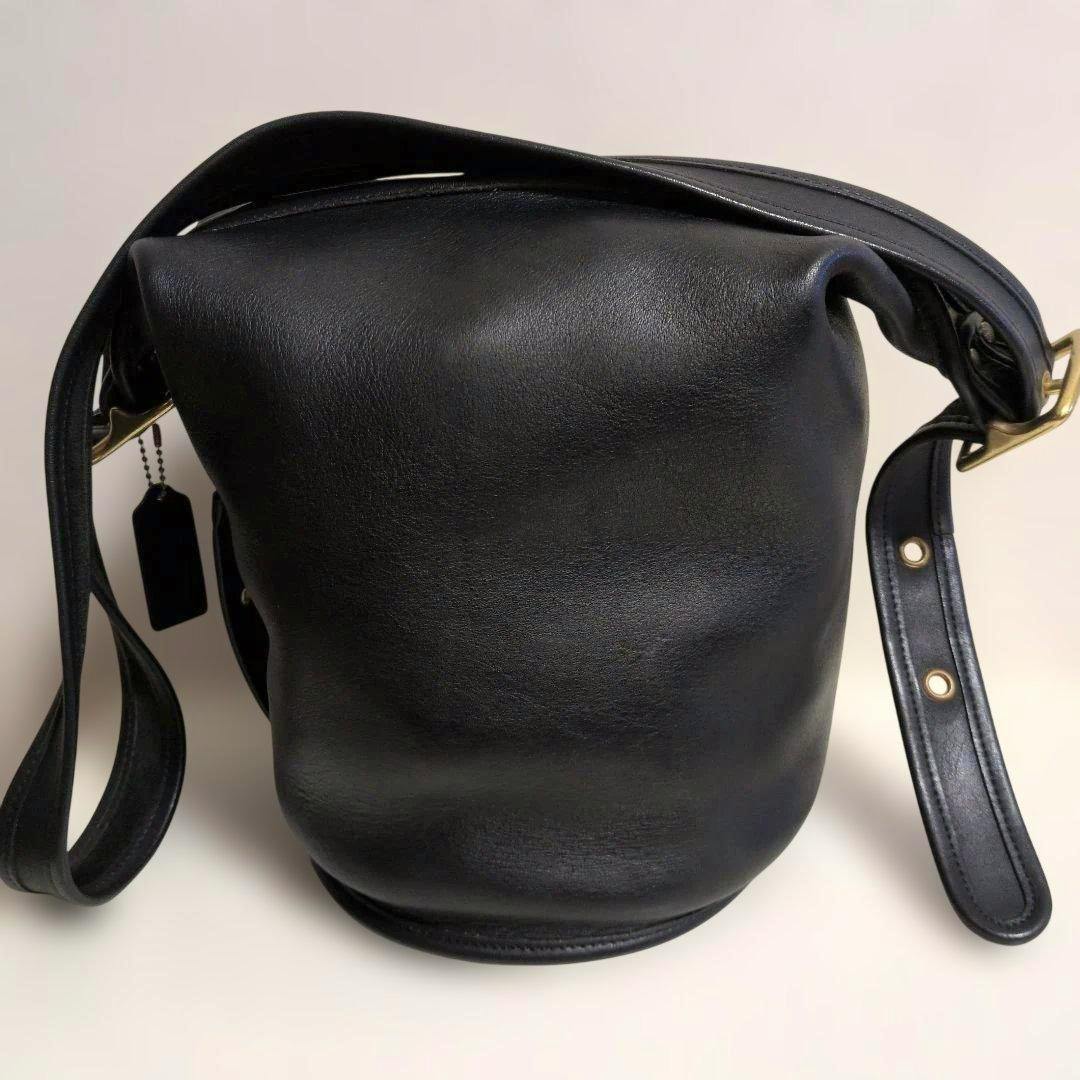 Front view of a black Coach bucket-style leather shoulder bag with an adjustable strap and a leather hang tag.