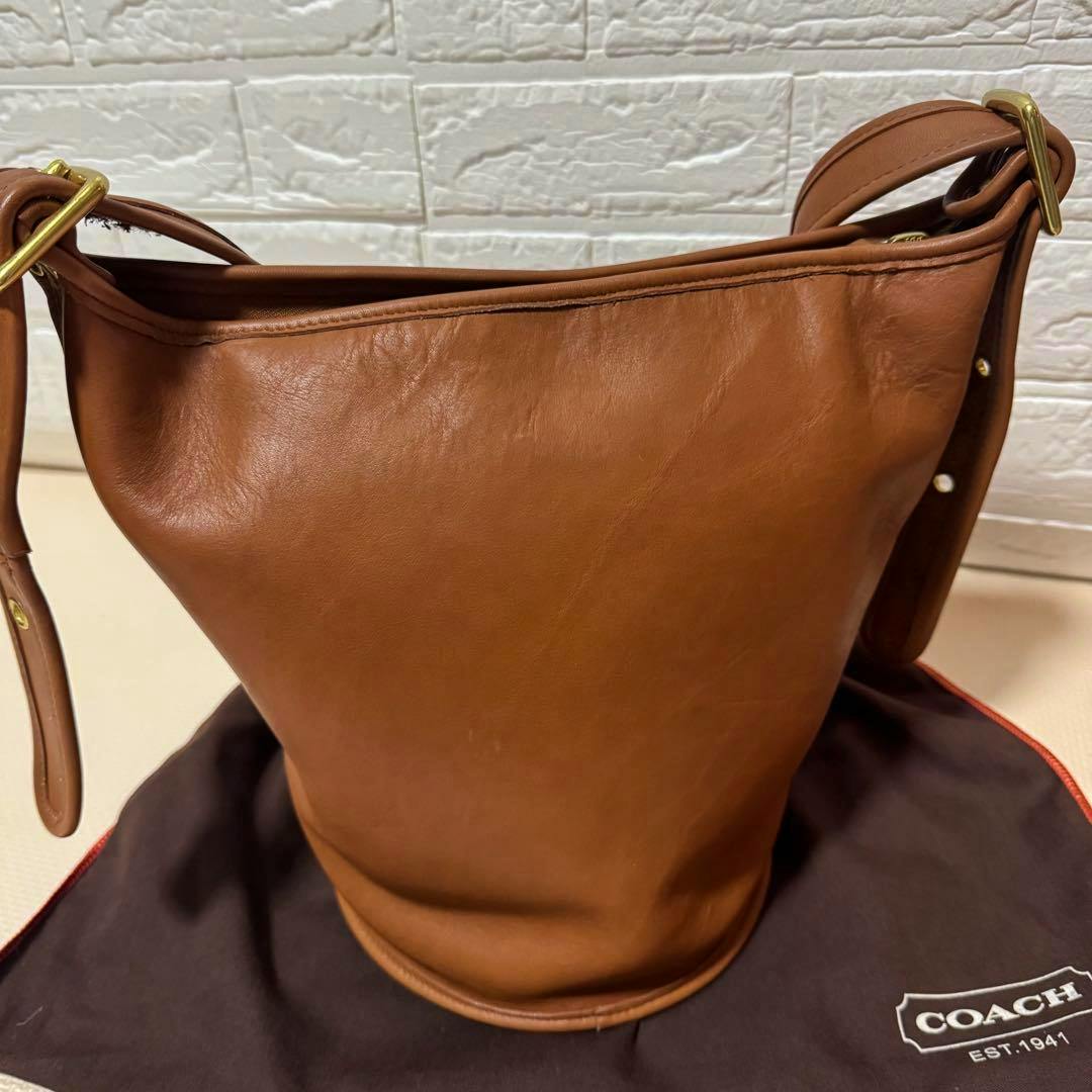 Brown Coach leather bucket-style shoulder bag standing upright with front and side visible on a Coach dust bag.