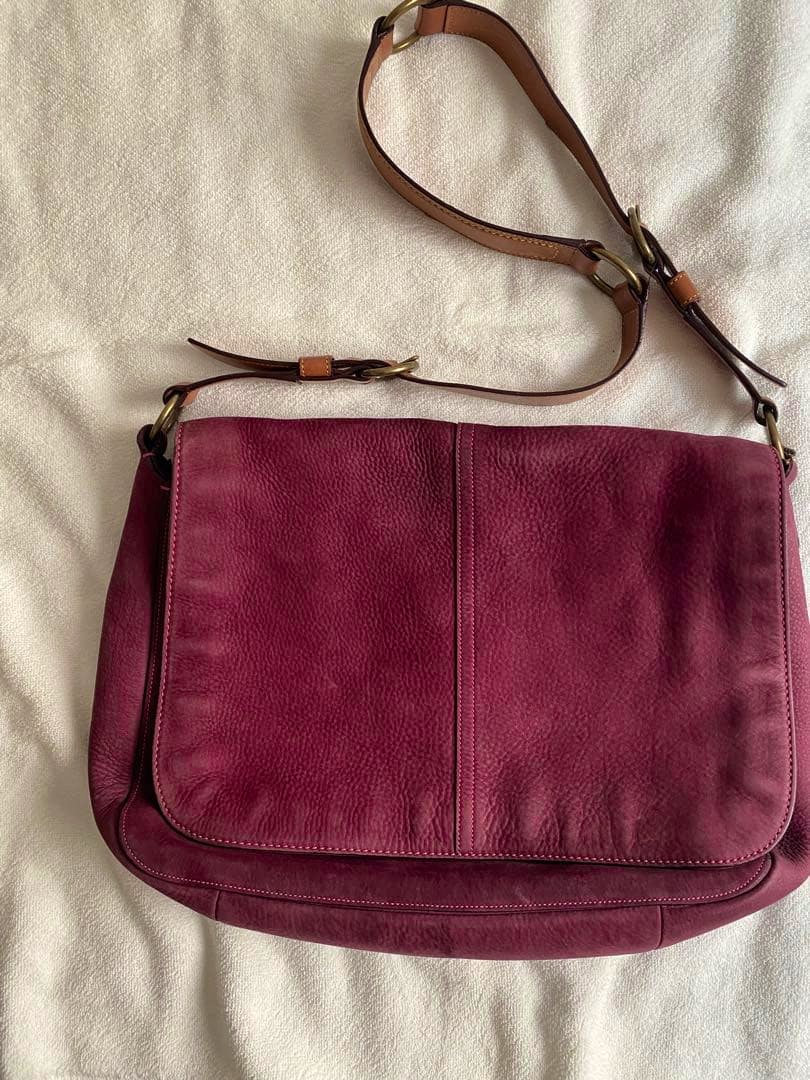 Front view of a burgundy Coach leather messenger bag with an adjustable brown shoulder strap on a light background.