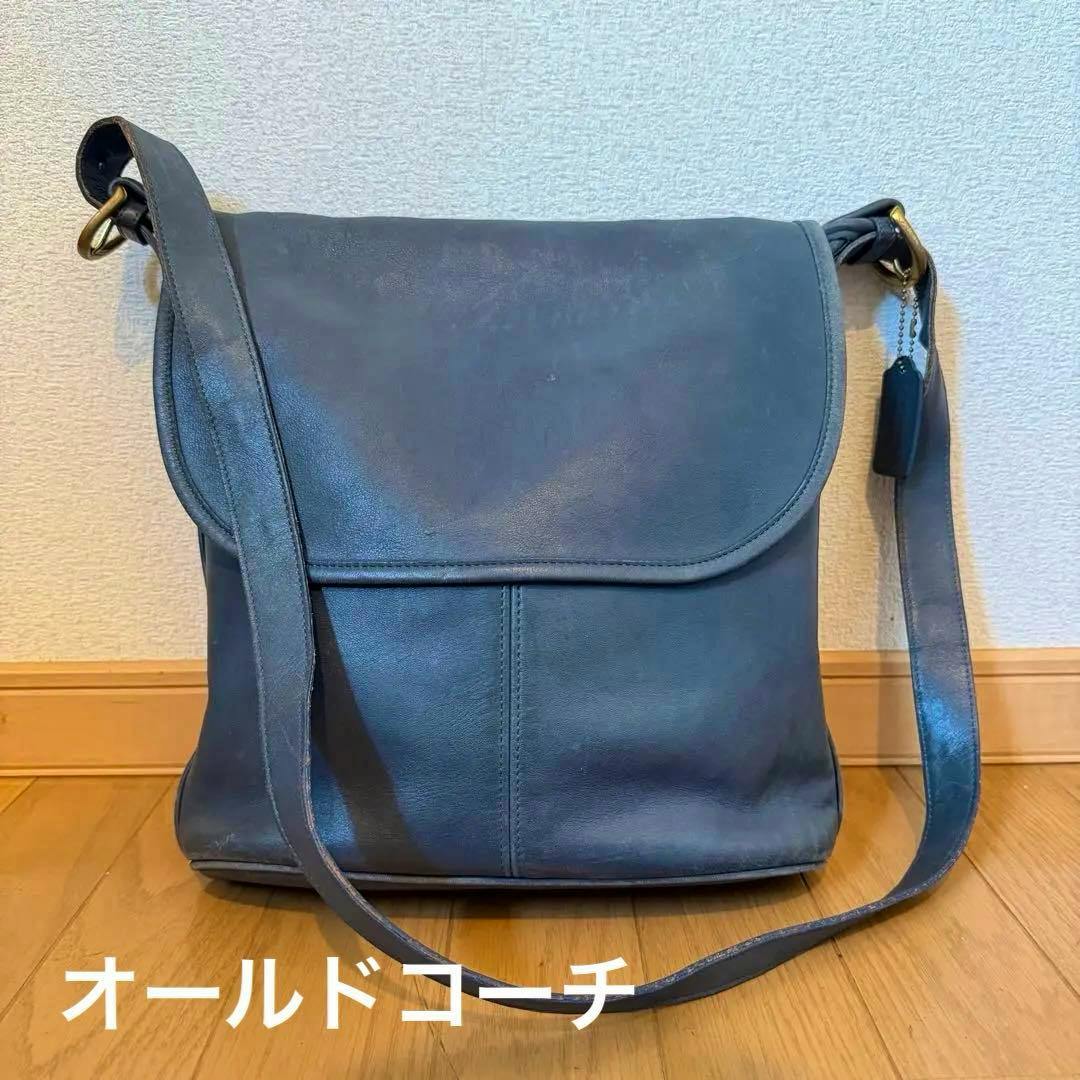 A dark cow-leather COACH shoulder bag with a flap and long strap standing on a wooden floor against a wall.