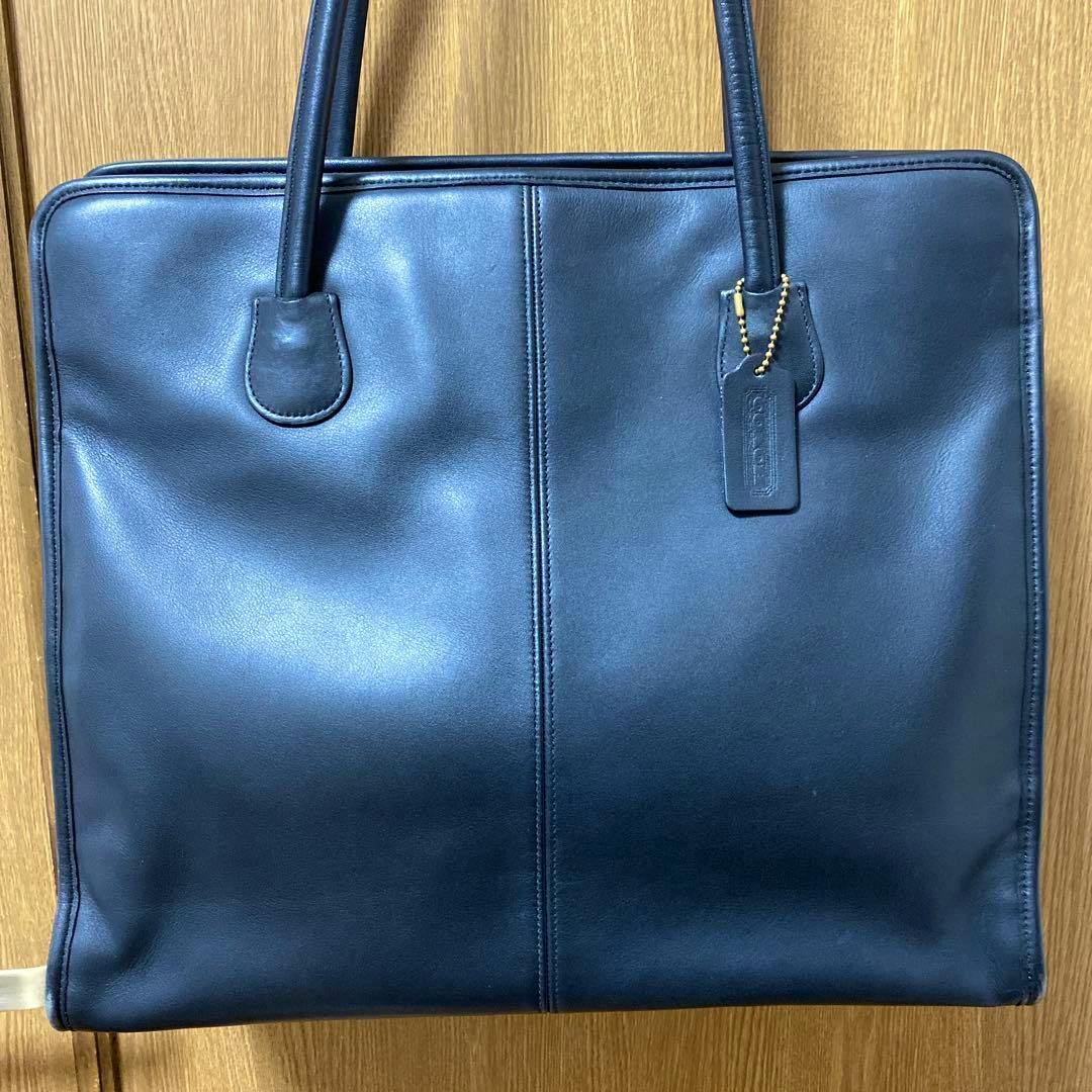 Front view of a black COACH leather tote bag with handles and a leather tag hanging on a wooden door.