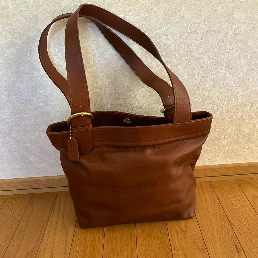 Brown COACH leather tote bag with double shoulder straps shown standing on a wooden floor against a light wall.