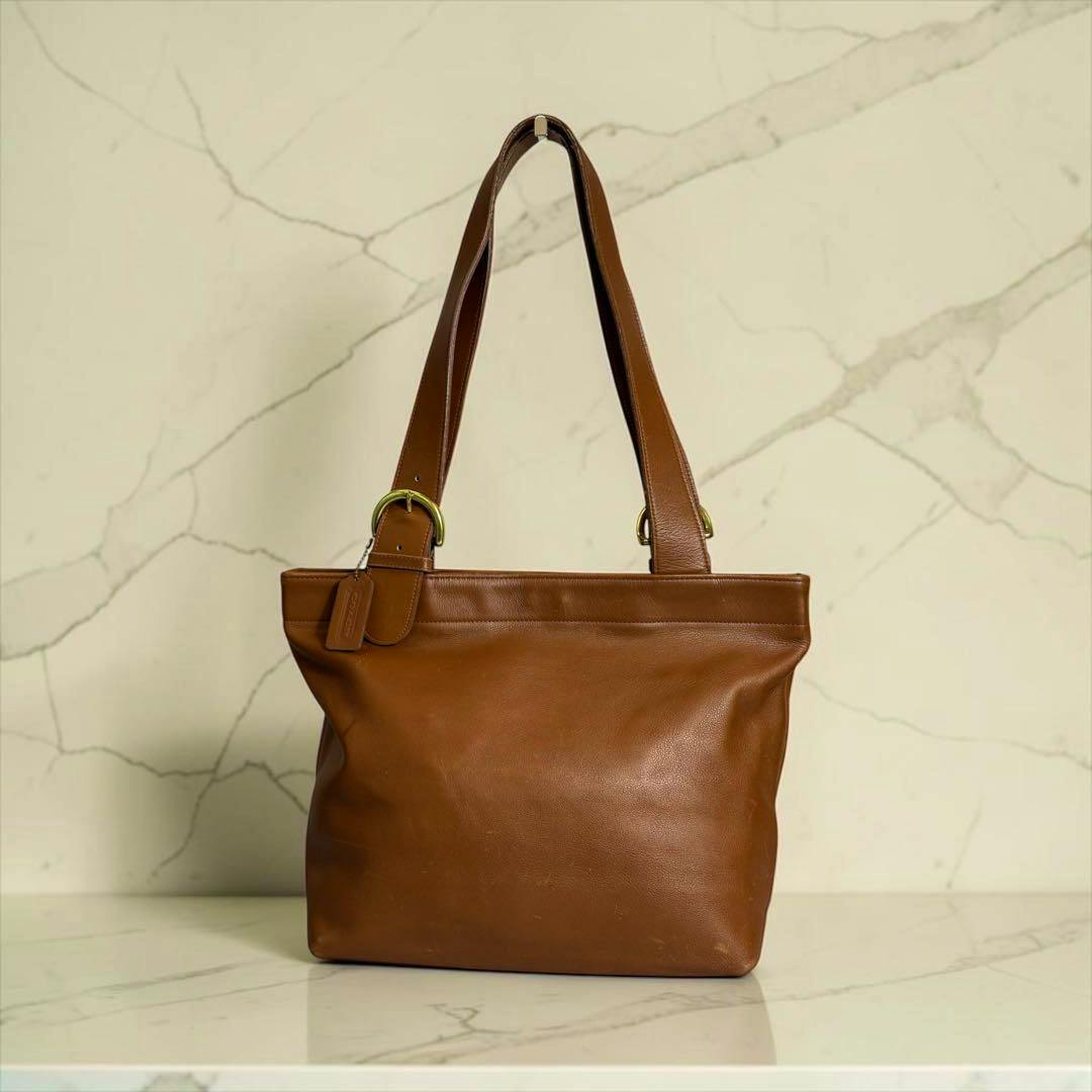 Brown leather Coach tote bag with shoulder straps shown against a light marble background.
