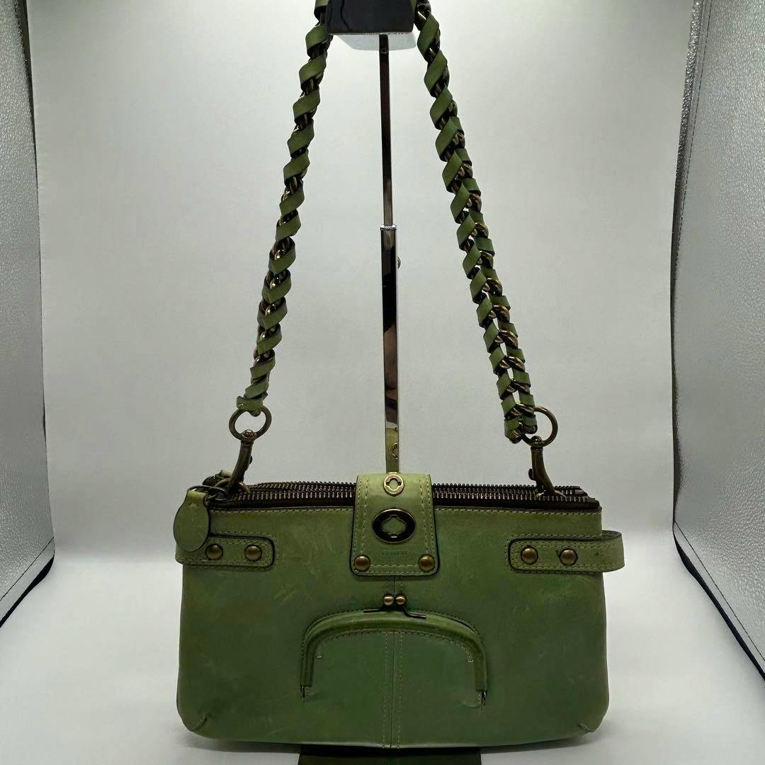 Green COACH Vintage Legacy turnlock leather shoulder bag with braided strap, front zip pocket, and brass-tone hardware displayed on a stand.