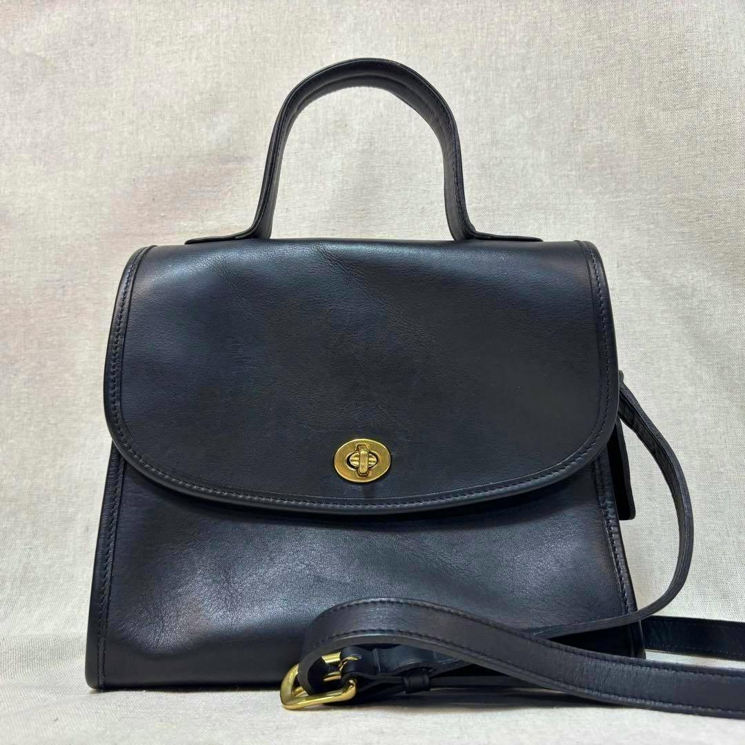 Front view of a black COACH Vintage Manor crossbody handbag showing the top handle, long strap, and gold turn-lock closure on a neutral background.