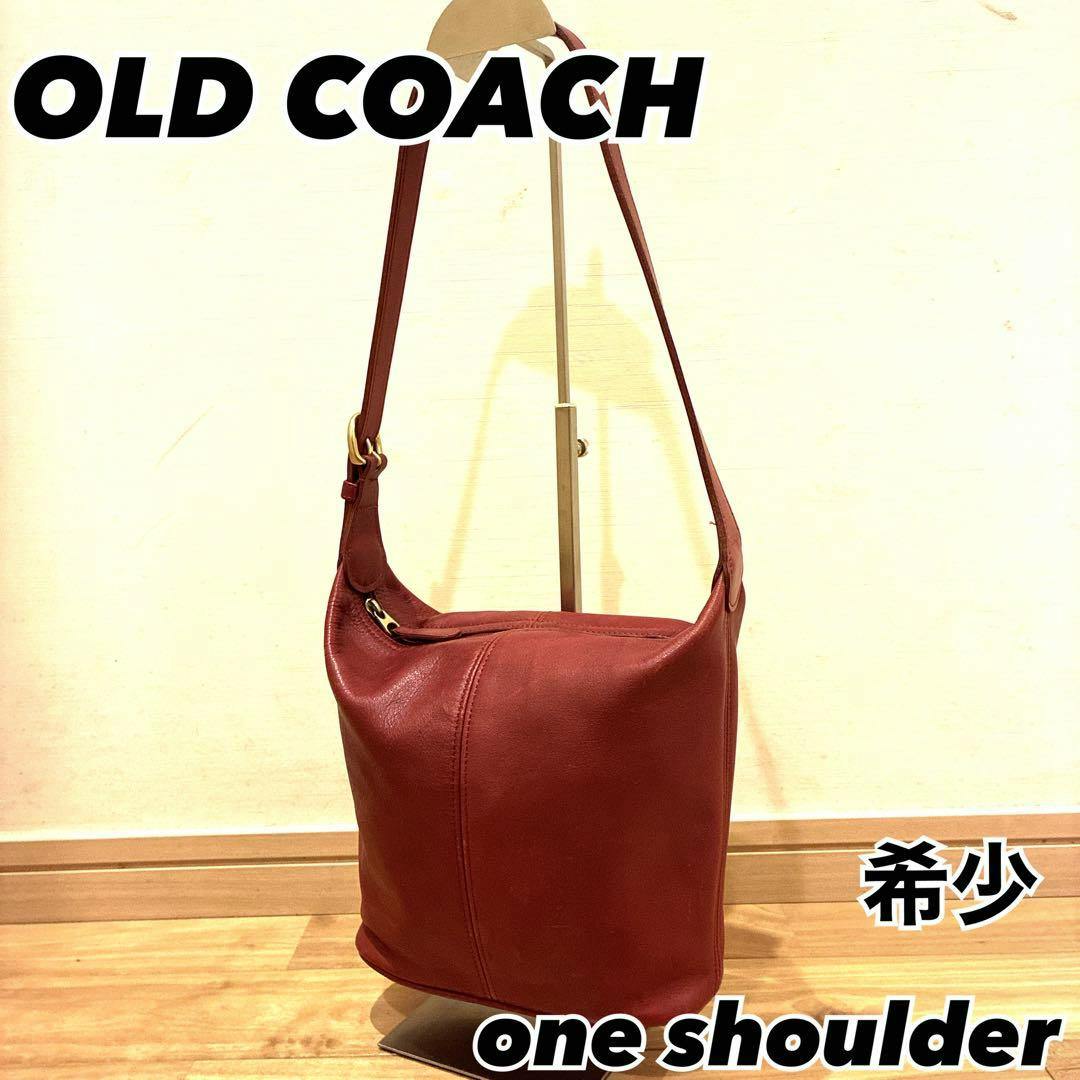 Red one-shoulder Coach leather bag hanging on a display stand against a plain wall.