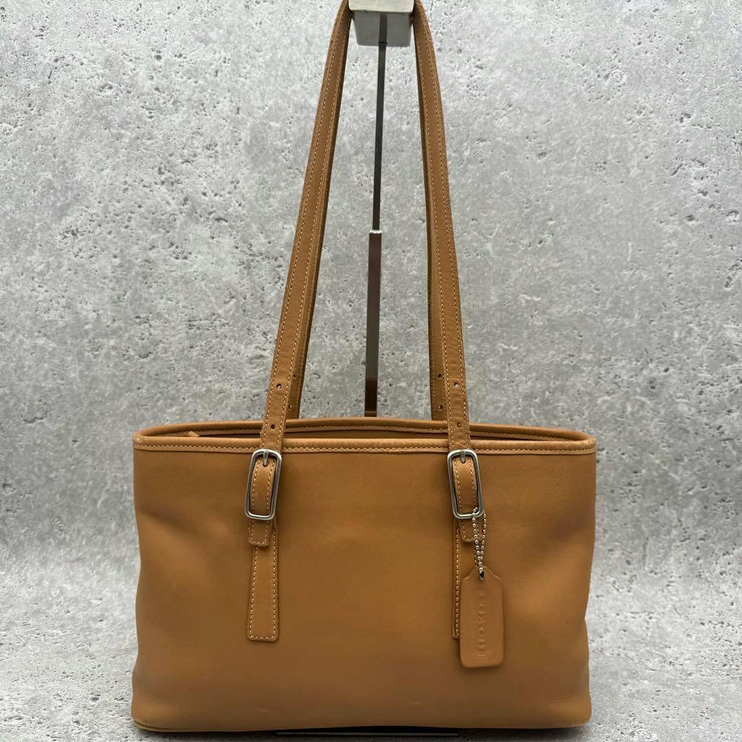 Camel COACH leather tote bag with two shoulder straps and a Coach hang tag shown against a gray background.
