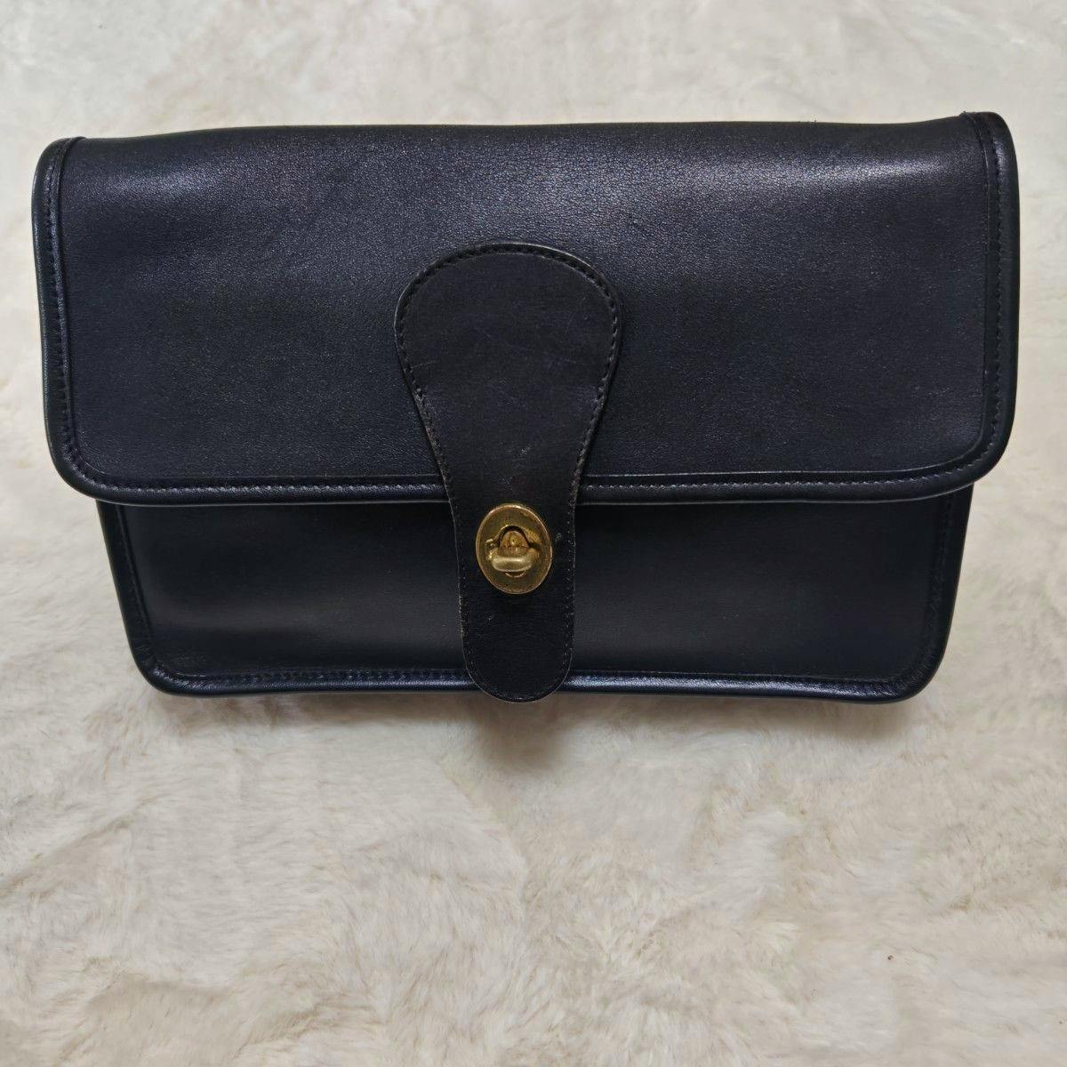 Front view of a COACH black leather clutch bag with a flap and a gold turn-lock closure.