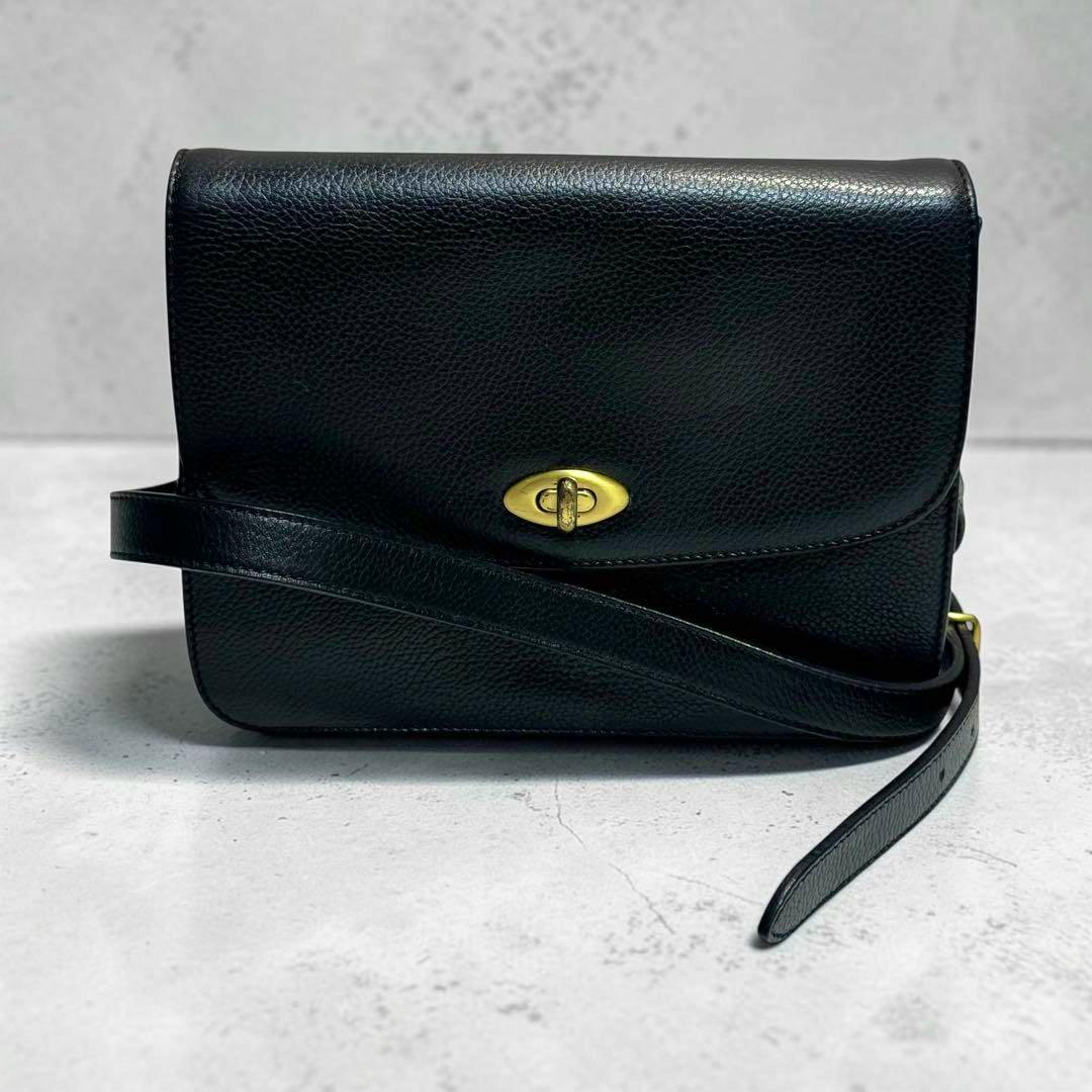Black Coach leather shoulder bag with a gold turn-lock closure and long strap shown on a neutral surface.