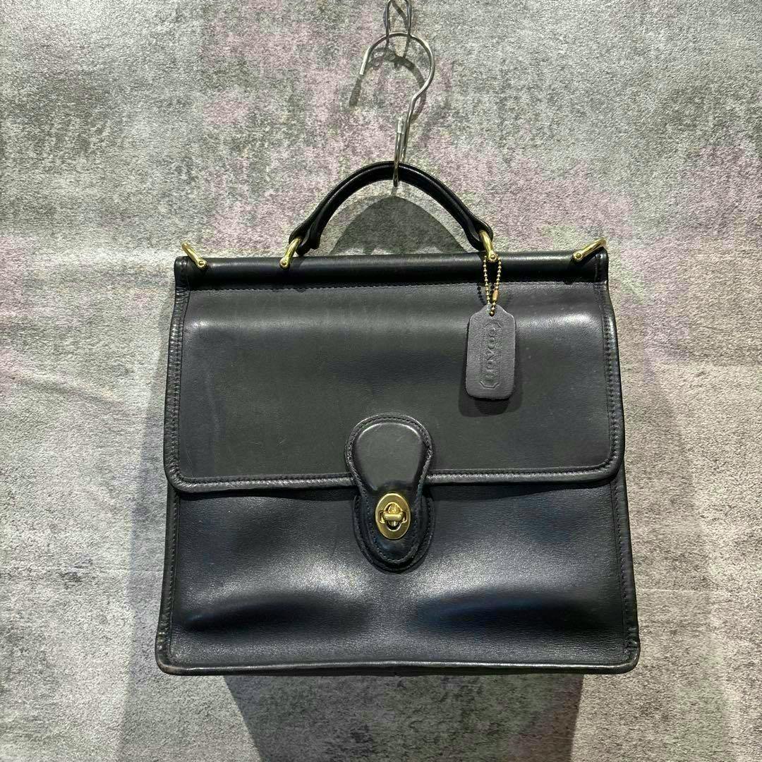 Black cow leather Coach vintage handbag with a top handle and gold turn-lock clasp hanging on a hook.