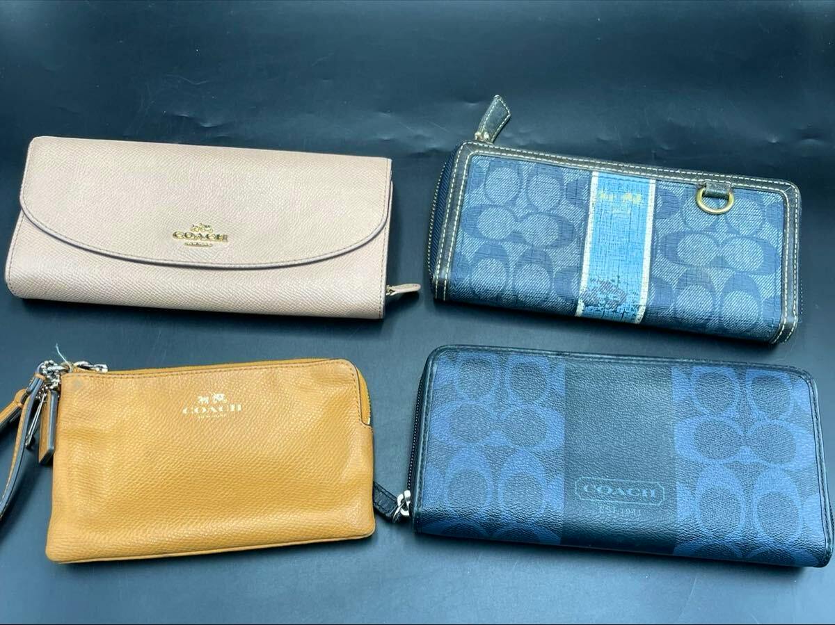 Four COACH wallets—a beige flap long wallet, a blue signature-pattern zip wallet with a stripe, a tan wristlet pouch, and a dark blue zip-around long wallet—arranged on a dark background.
