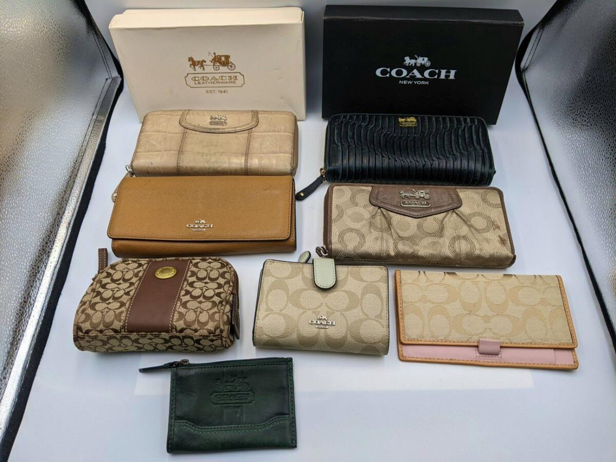 Eight Coach wallets and small pouches arranged on a white surface with two Coach boxes behind them.
