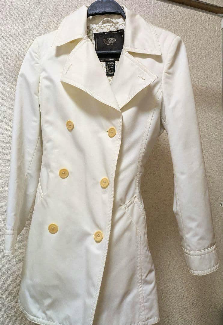 Front view of a white COACH trench coat on a hanger showing the double-breasted buttons and collar.