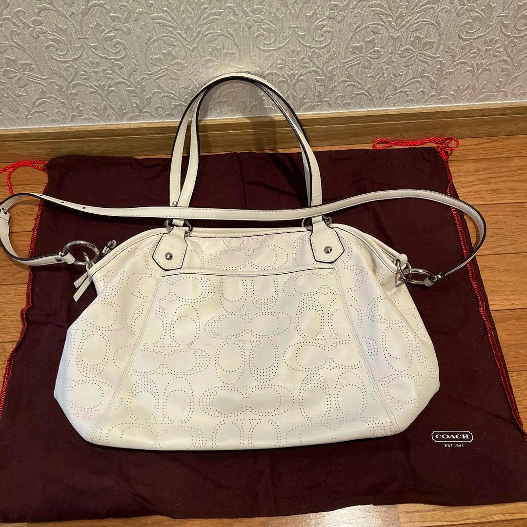 White COACH leather shoulder bag with a perforated pattern shown on a dark dust bag.