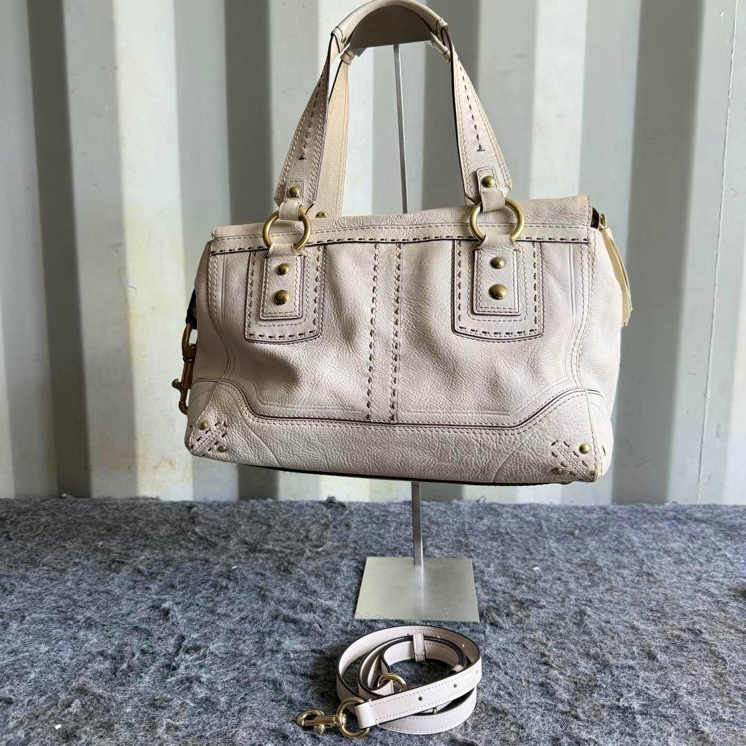 White leather Coach tote bag displayed on a stand with its detachable shoulder strap coiled on the surface below.