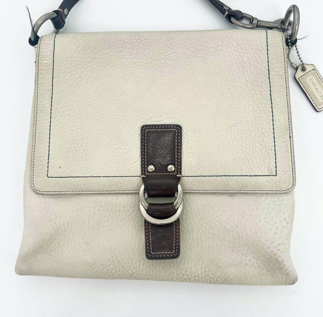 White synthetic pebbled shoulder bag with dark strap, front flap, metal ring clasp, and a Coach hang tag.