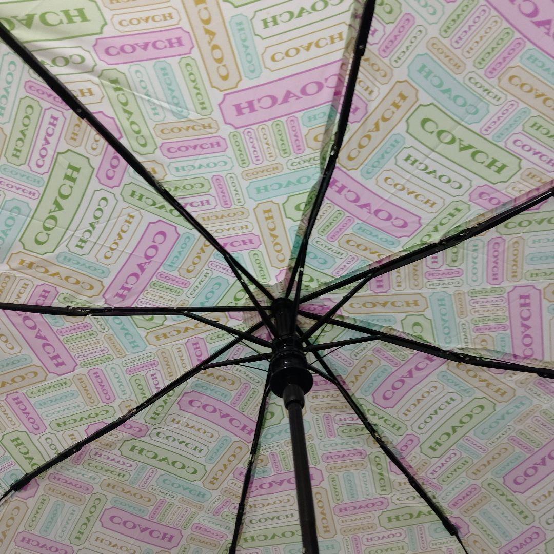 Interior view of an opened three-fold umbrella showing black ribs and a pastel canopy printed with COACH logos.