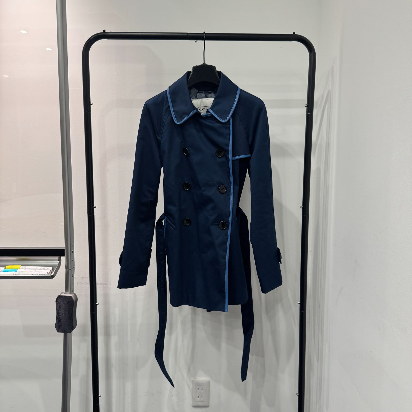 Navy women's cotton Coach trench coat with light contrasting piping hanging on a clothing rack against a white wall.