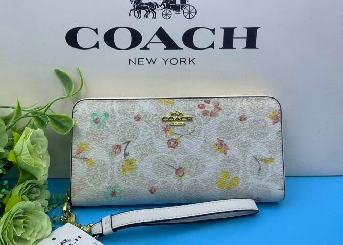 Front view of a white Coach long zip wallet with a floral print, gold logo plaque, and detachable wrist strap placed against a Coach box.
