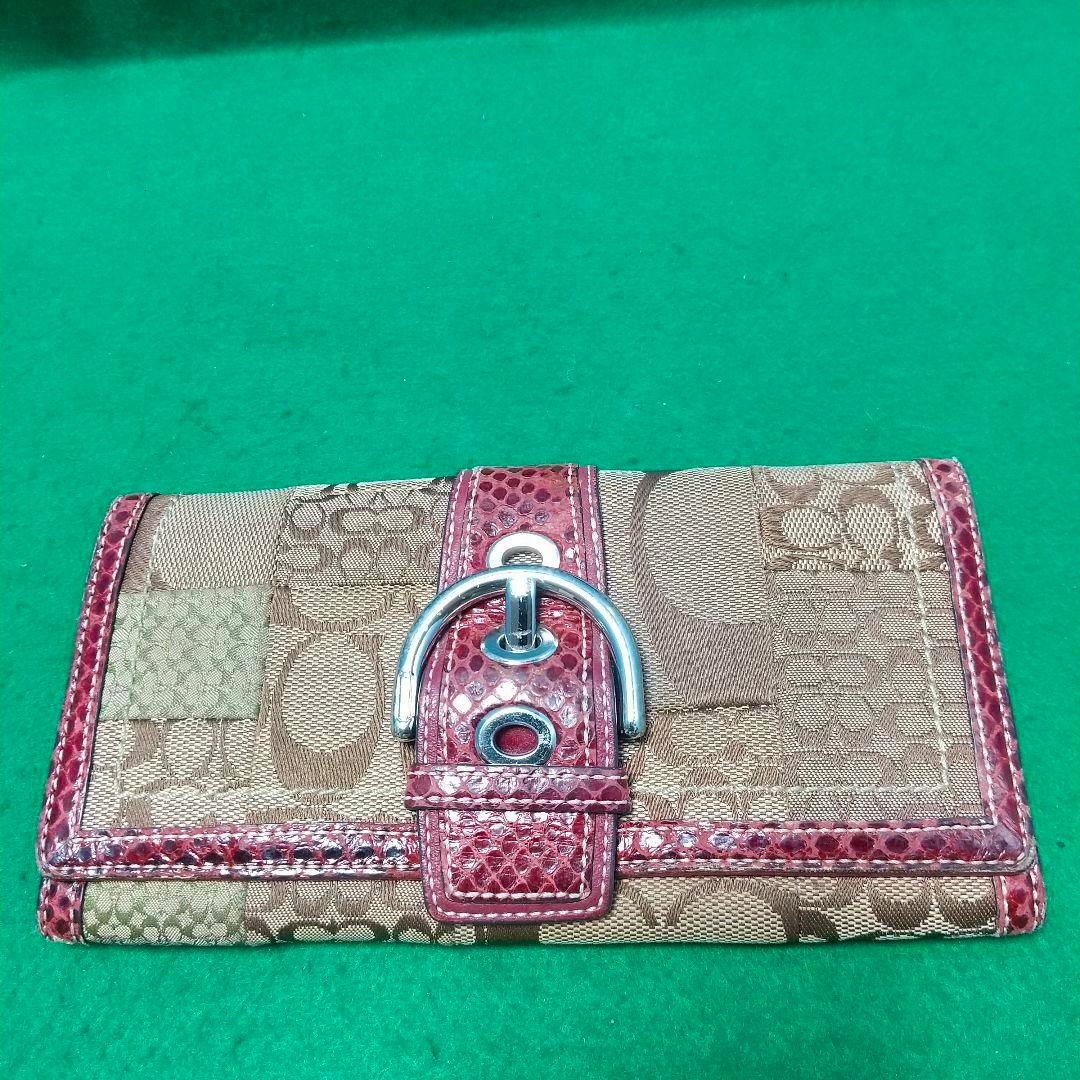 Front view of the Coach women's bi-fold long canvas wallet with signature print and a red belt-style strap with silver buckle on a green background.