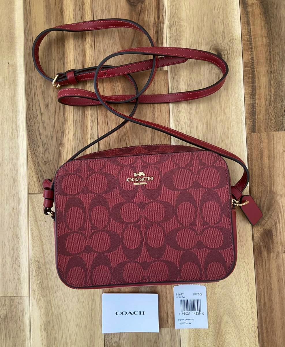 Front view of a red Coach signature shoulder bag with adjustable strap and tags on a wooden surface.