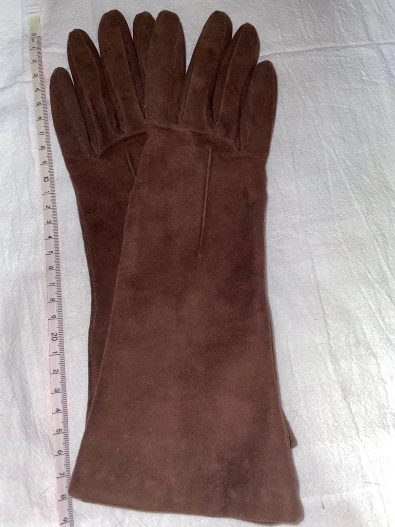 Pair of brown suede leather women's gloves laid flat on a cloth background with a measuring ruler beside them.