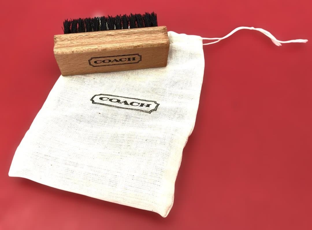 Wooden COACH cleaning brush with bristles resting on a white COACH storage pouch against a red background.