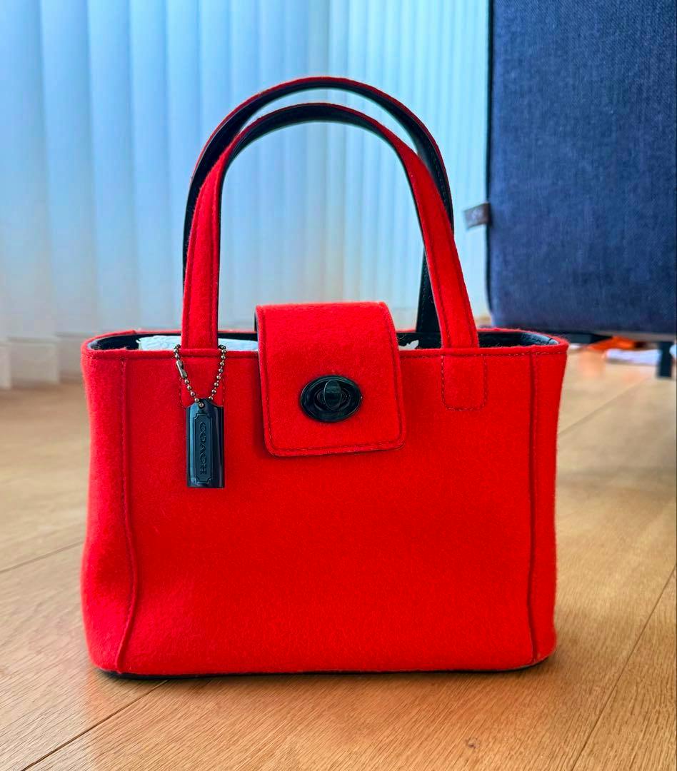 Red COACH wool felt tote bag shown from the front on a wooden floor with handles and a flap closure visible.