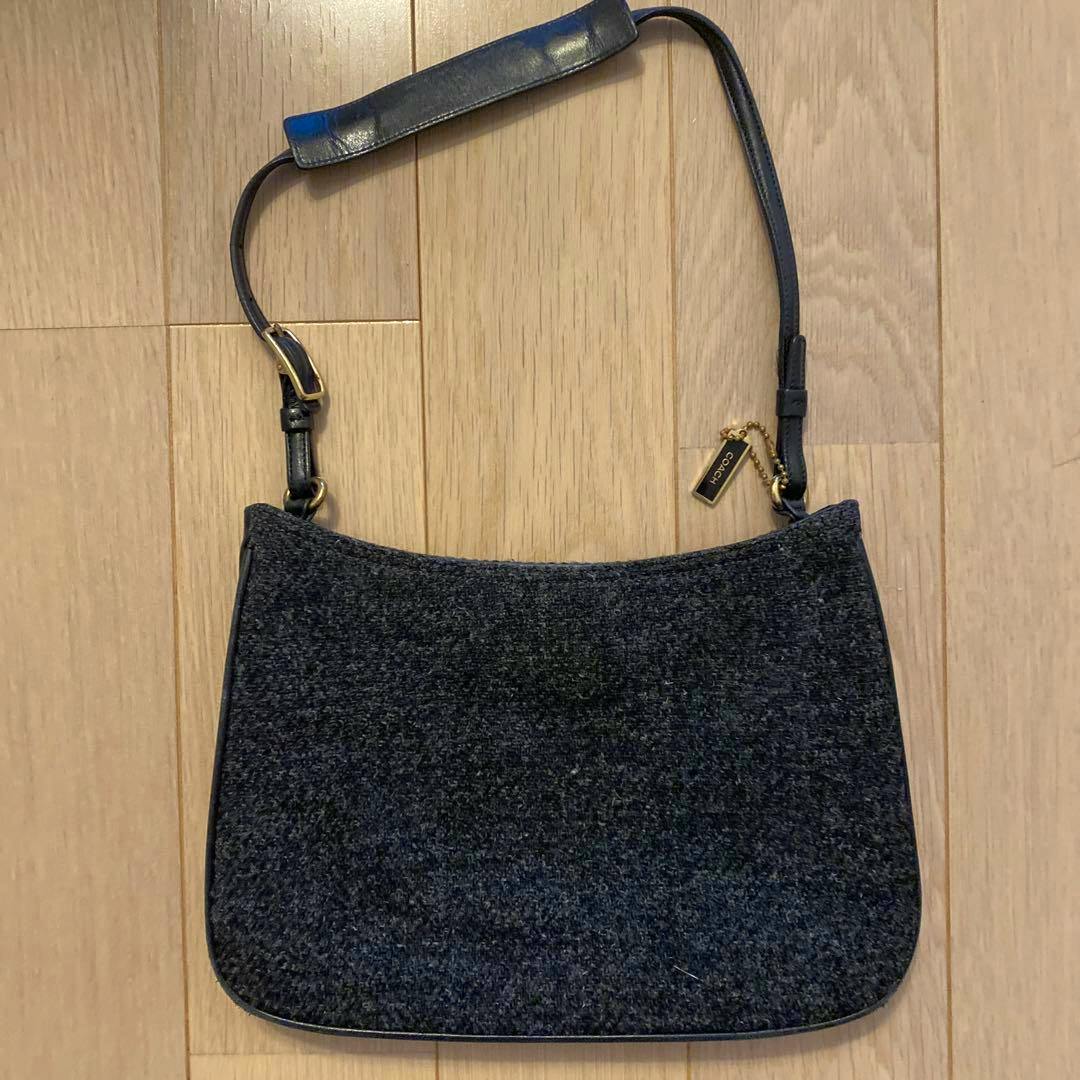 A black Coach wool shoulder bag shown flat on a wooden floor, with the front and strap visible.