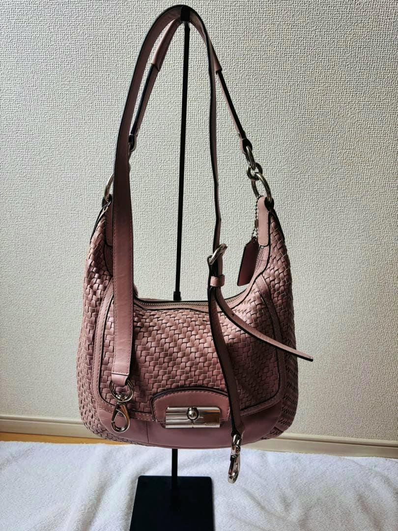 Pink COACH woven leather shoulder bag hanging on a display stand against a wall.