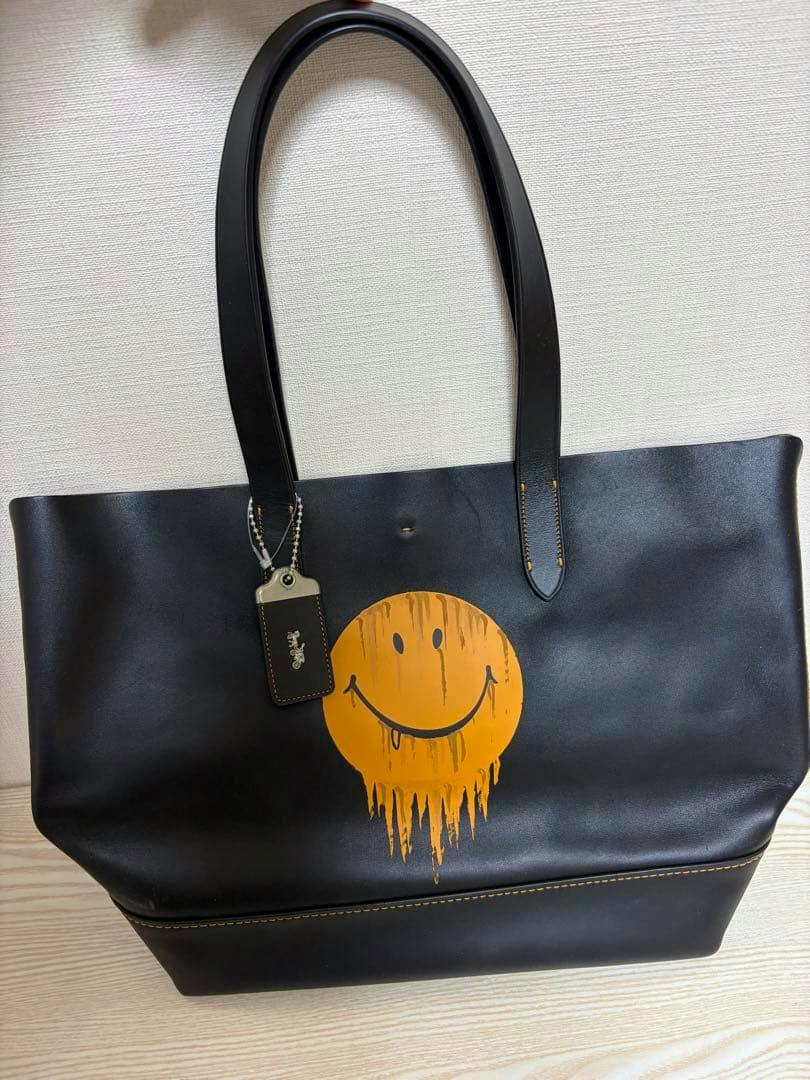 Black COACH×BASEMAN tote bag with long handles and a dripping yellow smiley-face graphic on the front.