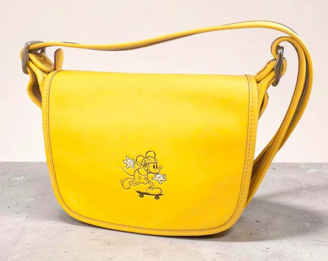 Front view of a yellow leather COACH and Disney limited-collaboration shoulder bag showing a small Disney character graphic on the front.