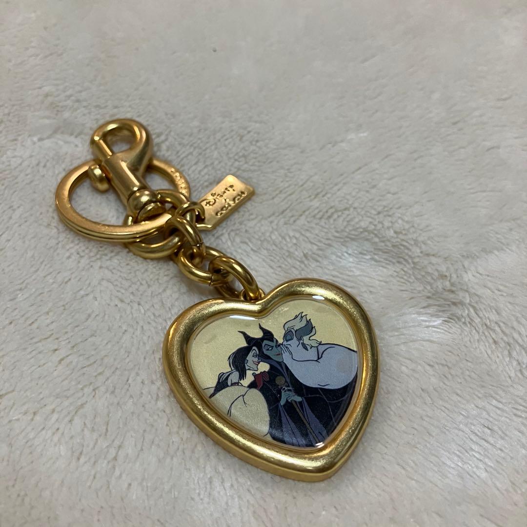 Gold-tone heart-shaped COACH x Disney Villains keychain charm showing printed villain artwork in the center.