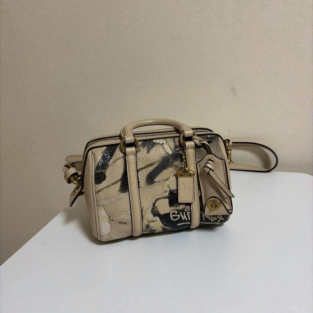 Cream leather COACH x MINT + SERF Ruby Satchel 18 on a white surface showing the front with graffiti-style print, top handles, shoulder strap, and hanging tag.