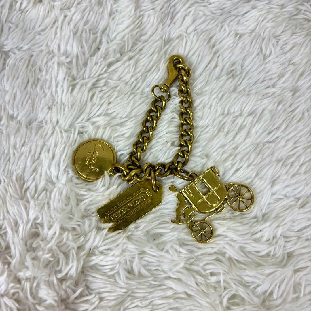 Gold Coach horse carriage bag charm keychain with chain, rectangular tag, and round logo charm on a white fuzzy background.