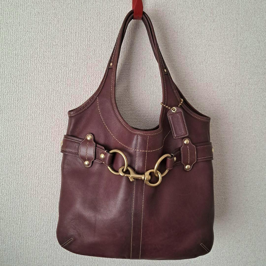 Front view of a brown Coach cow-leather hobo handbag with gold horsebit hardware hanging on a wall.