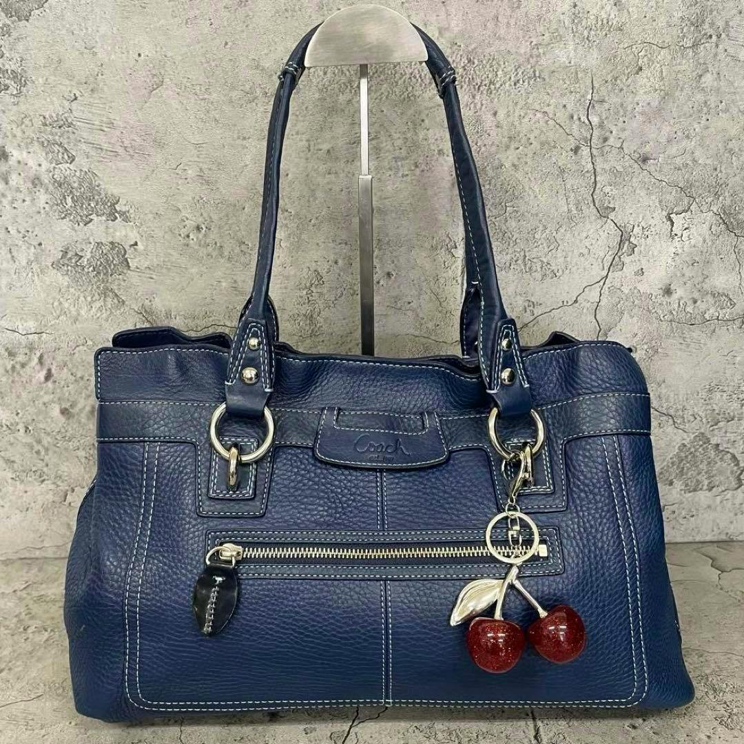 Front view of a navy COACH leather Boston tote bag with double handles, a front zip pocket, contrast stitching, and a cherry key charm.