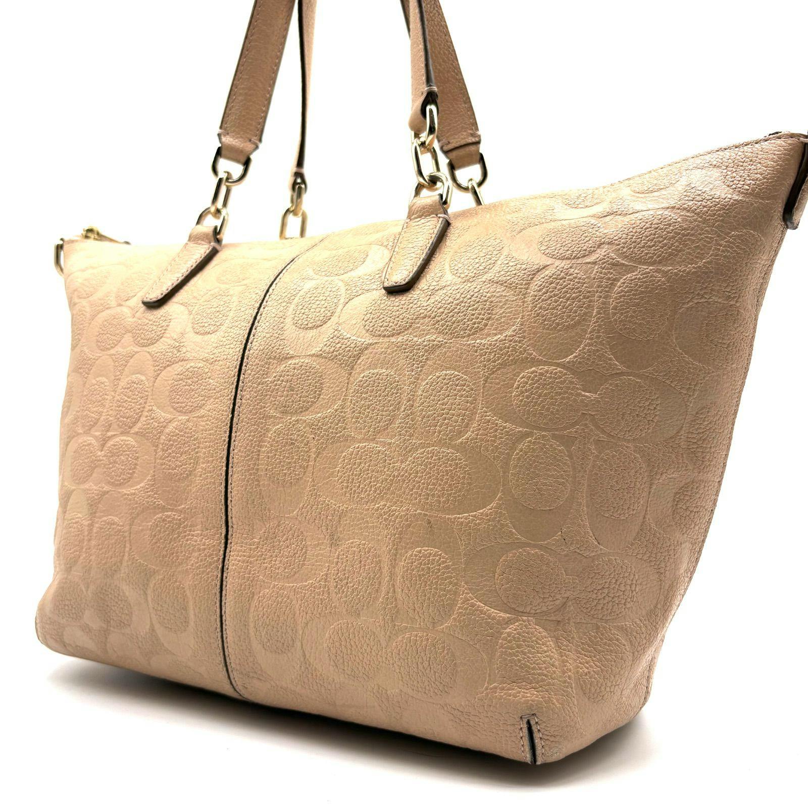 Close-up of a beige COACH embossed leather satchel bag showing signature pattern and top handles.